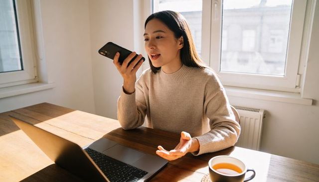 Woman speaking into smartphone while working remotely with laptop and coffee