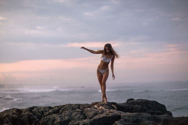 A woman wearing a bikini is balancing on rocks along the shore at sunset. The ocean and a colorful horizon are in the background. Perfect for use in travel, leisure, lifestyle, beachwear promotion, relaxation or nature-related content.
