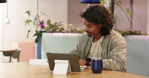 African American man taking notes on tablet in pastel coworking cafe with tumbler