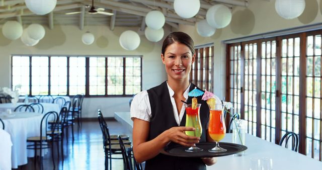 Female server with tray holding two colorful drinks, standing in an empty and elegantly decorated restaurant. Suitable for concepts related to customer service, hospitality industry, dining, and restaurant marketing. Ideal for advertisements, brochures, and articles emphasizing professional service and quality hospitality.