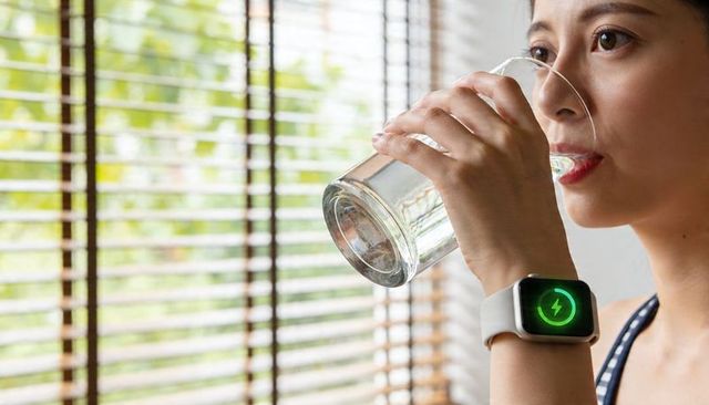 Woman drinking water while smartwatch showing charging icon on wrist by window blinds