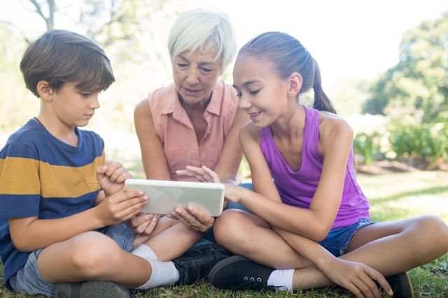 Grandmother and her grandchildren are sitting on the grass in a park on a sunny day, using a digital tablet together. This image can be used to depict family bonding, intergenerational learning, and the use of technology in everyday life. It is ideal for advertisements, educational materials, and articles about family, technology, and outdoor activities.