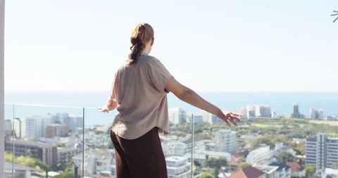 Gazing woman on high-rise balcony overlooking coastal cityscape and ocean horizon