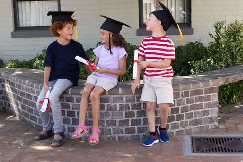 Children celebrating kindergarten graduation on brick wall wearing caps holding diplomas