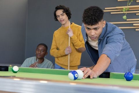 Diverse young men lining up precise pool shot in modern recreation lounge with green felt