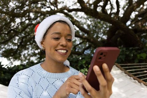 African american woman wearing santa hat smiling while using smartphone in backyard hammock