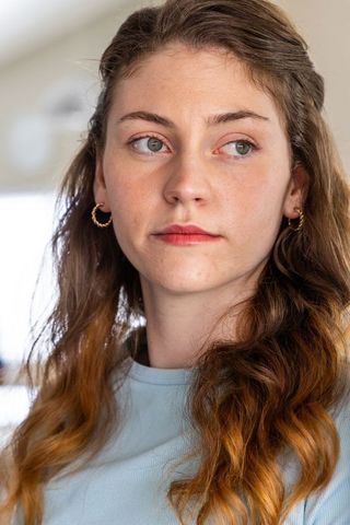 Introspective young woman gazing left by window wearing twisted gold hoop earrings, blue top