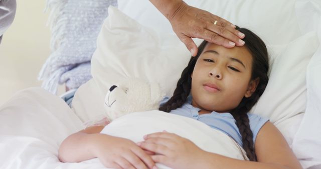 Girl lying in bed with eyes closed, holding plush toy, while adult hand touches her forehead to check for fever. Ideal for topics on childhood illness, healthcare, parenting, parental love, and home care strategies.