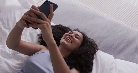 Smiling african american woman lying on bed using smartphone and relaxing in blue camisole