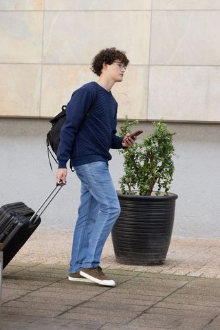 Young man walking with rolling suitcase and checking smartphone beside urban tiled wall