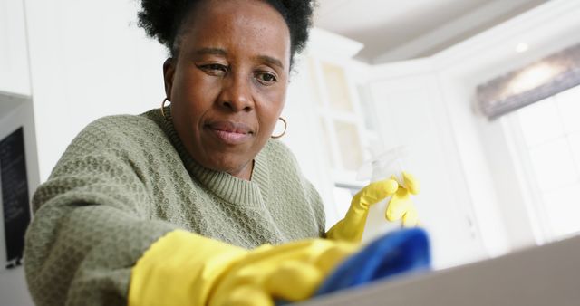 An African American woman is cleaning the kitchen counter with a focused expression. She is wearing yellow cleaning gloves and a green sweater, diligently ensuring cleanliness. This image can be used for topics relating to hygiene, cleanliness, housekeeping, domestic cleaning services, and sanitation in the home.