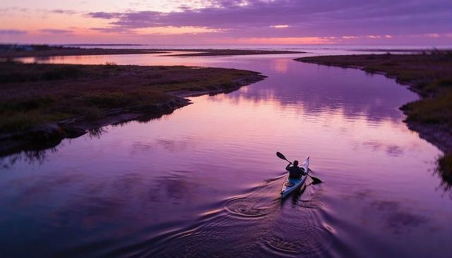Solo kayaker paddling through purple estuary at sunset, marsh channel and ripples