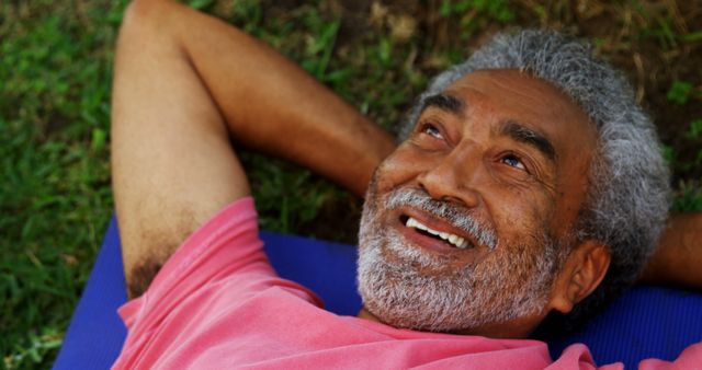 Elderly man with grey hair, lying down on grass in outdoor park, smiling. Ideal for advertisements, health and wellness promotions, lifestyle blogs, and senior living content. Shows relaxation, happiness, and leisure for senior demographic.