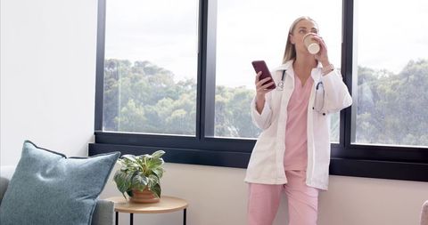 Female nurse in pink scrubs leaning at window sipping coffee and checking smartphone