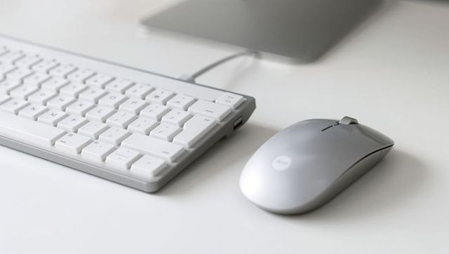 Silver wireless mouse on white desk with slim keyboard and laptop, minimalist workspace