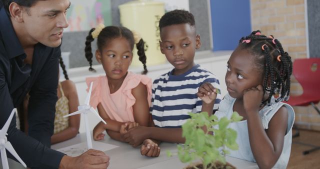 Teacher presenting to curious children while demonstrating wind turbines model in classroom. Group of young learners surrounded by clean energy projects, engaged in interactive learning environment. Ideal for education, STEM topics, renewable energy awareness, and diversity in learning settings.