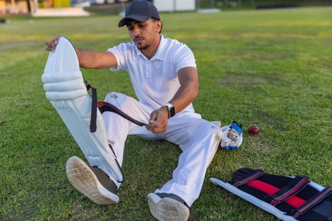Indian cricketer fastening batting pads on sunlit field wearing white kit and black cap