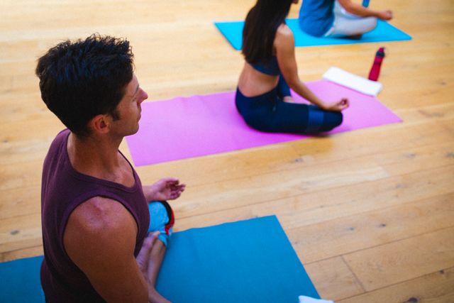 Young adults sitting on yoga mats meditating in a yoga studio. Ideal for promoting fitness, wellness, and healthy lifestyle programs. Suitable for use in articles, blogs, and advertisements related to yoga, meditation, and group exercise classes.