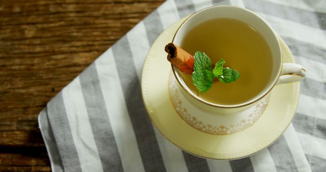 A cup of tea with a cinnamon stick and mint leaf garnish sits atop a saucer on a striped napkin, with copy space. The warm beverage provides a sense of comfort and relaxation, perfect for a cozy break.