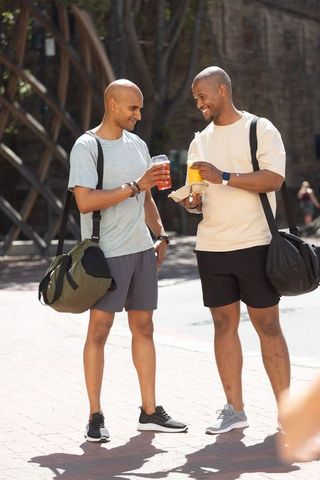 African american men chatting after workout, carrying duffel bags and drinks in sunlit plaza
