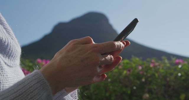 Hands holding smartphone during sunny day with mountain in background. Great visual for topics related to technology, travel, outdoor activities, communication in nature, and connectivity. Perfect for apps and websites promoting travel tips, mobile technology, and scenic vacation spots.