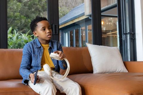 African american boy holding headphones sitting on modern tan sofa wearing denim jacket