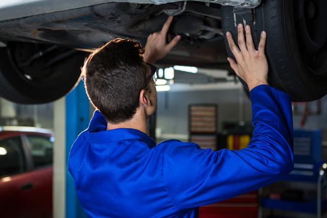 Mechanic in blue uniform inspecting car undercarriage in a repair garage. Ideal for use in automotive service advertisements, repair shop promotions, and articles on car maintenance and professional mechanics.