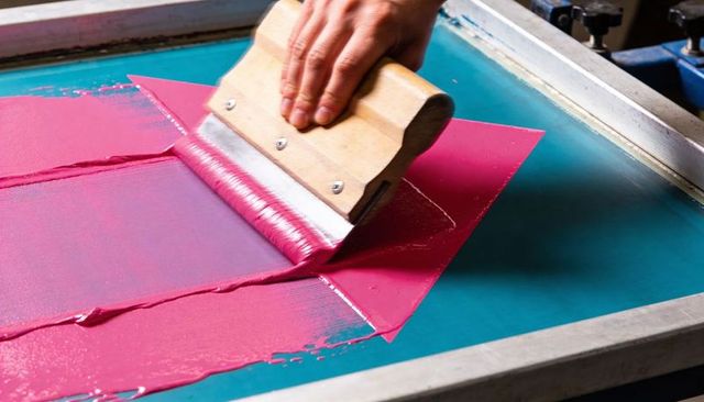 Hand pulling magenta ink with wooden squeegee on turquoise screen printing frame