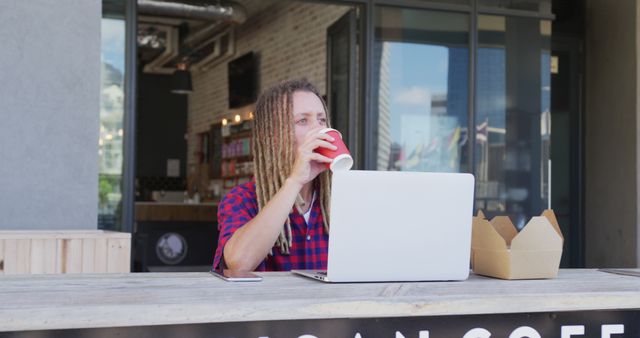 Man with dreadlocks enjoys coffee while working on laptop at outdoor table of a modern cafe. The casual and urban setting is perfect for portraying remote work, digital nomad lifestyle, or coffee culture. Ideal for use in articles or advertisements focused on freelance work environments, outdoor dining experiences, or modern urban living.