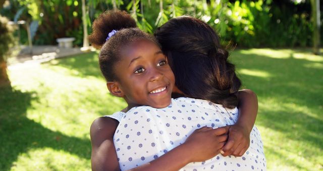 This heartwarming image captures a happy African American girl with puffy hair, hugging her mother outdoors in a lush green garden. Ideal for use in family, parenting, and outdoor activities promotional materials. It conveys warmth, love, and bonding in a family setting. Suitable for articles, advertisements, and brochures focused on familial relationships and outdoor leisure.