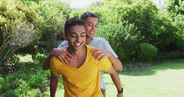 Smiling biracial gay male couple having fun piggybacking in garden. staying at home in isolation during quarantine lockdown.