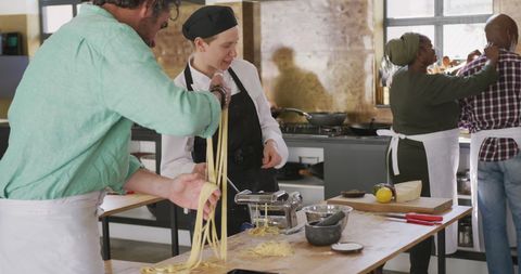 Diverse group participates in a hands-on cooking class focused on making fresh pasta in a modern kitchen. Center of activity shows experienced female cook in chef hat guiding a senior man with pasta preparation tools. Lemon, parmesan, and other ingredients highlight a practical culinary environment. Perfect for illustrating educational food experiences, promoting culinary workshops, or teaching team-building exercises through cooking.