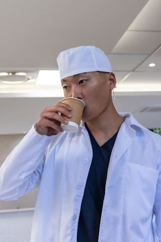 Korean physician wearing lab coat and surgical cap drinking coffee during clinic break