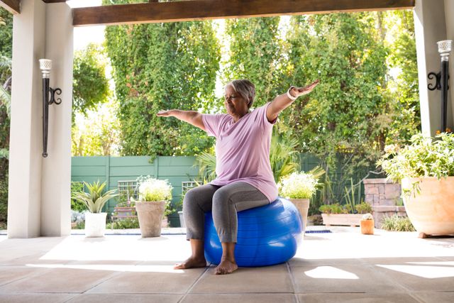 Senior woman with arms outstretched sitting on exercise ball under shed in yard