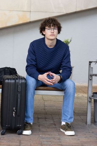 Curly-haired young man sitting on bench holding smartphone next to rolling suitcase