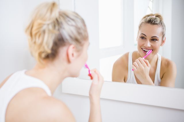 Young woman brushing teeth in front of bathroom mirror, promoting good dental hygiene and healthy morning routines. Ideal for use in health and wellness blogs, dental care advertisements, personal care products, and lifestyle magazines.