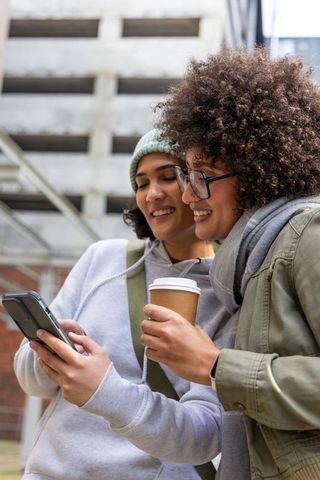 Two friends checking smartphone while holding coffee near urban parking garage