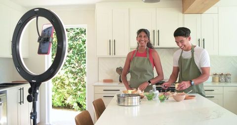 Multicultural couple recording cooking tutorial using ring light and smartphone in bright kitchen