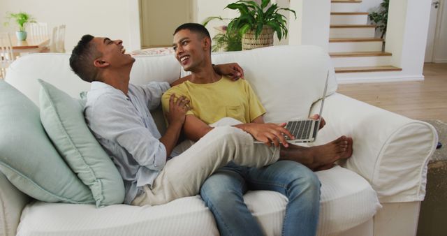 Young gay couple spending time together on a couch at home. They are laughing and seem happy and affectionate while one partner holds a laptop. Ideal for visuals related to LGBTQ+ relationships, love, home settings, and modern living.