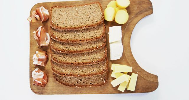 Slices of whole grain bread are neatly arranged on a wooden cutting board next to various types of cheese and cured meats, with copy space. A simple yet inviting setup for a rustic sandwich or charcuterie board.