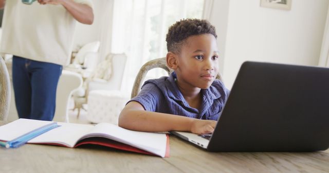 A young boy is seated at a table, using a laptop for his homework. The scene is set in a bright and comfortable home. An open notebook and some pencils are also on the table. This image is perfect for illustrating remote learning, homeschooling, or educational technology in everyday life.