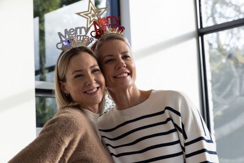 Two women wearing merry christmas headbands leaning heads and smiling in bright home