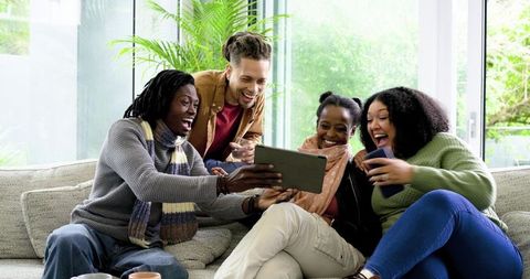 Group of friends laughing and sharing tablet and smartphone on cozy living room sofa