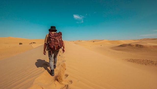 Walking lone hiker along dune crest with leather backpack and brimmed hat under blue sky