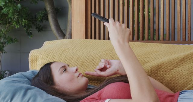 This image features a young woman lying on a couch holding and looking at her smartphone. She appears relaxed in a casual indoor setting with a yellow cushion behind her. Useful for illustrating concepts like home leisure, digital lifestyle, youth, relaxation, and technology use at home. Ideal for blogs, articles, and advertisements focusing on modern technology and everyday life.