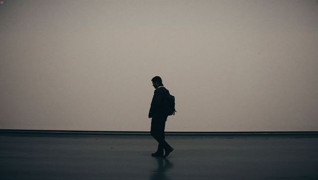 Solitary man walking with backpack in dim minimalist gallery corridor with reflection