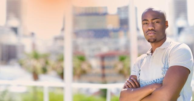 Young black man standing outdoors with arms crossed against a backdrop of city buildings. The urban landscape in the background is blurred, emphasizing the subject's confidence and modern style. This image can be used for promoting urban fashion, lifestyle blogs, and confidence in business settings.