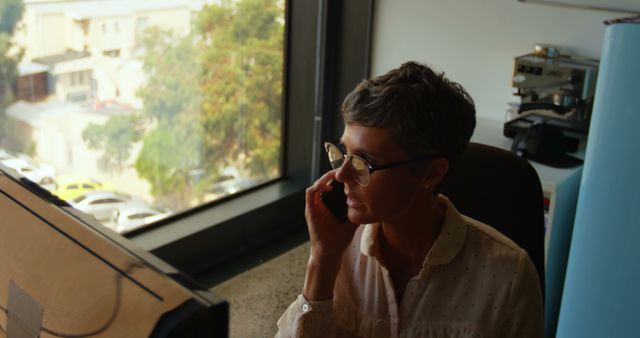 Businesswoman with short hair is having a professional phone call while sitting at a desk in an office setting. Ideal for conveying themes of communication, business, professionalism, and office work. Can be used in business blogs, professional websites, and corporate materials to represent women in leadership roles.