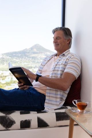 Serene mature man relaxing and reading novel on window seat with mountain view and tea