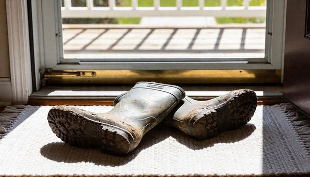 Resting muddy green rubber boots on woven doormat at sunlit open doorway